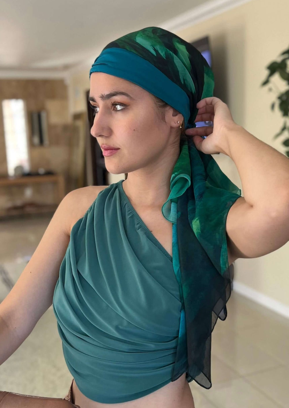 Beautiful lady in a luxurious chiffon and silk headscarf. The abstract print is peacock colored with brush strokes of black and green. her hand is on the back of her head and she has a sexy one shoulder top on, that is also peacock colored.